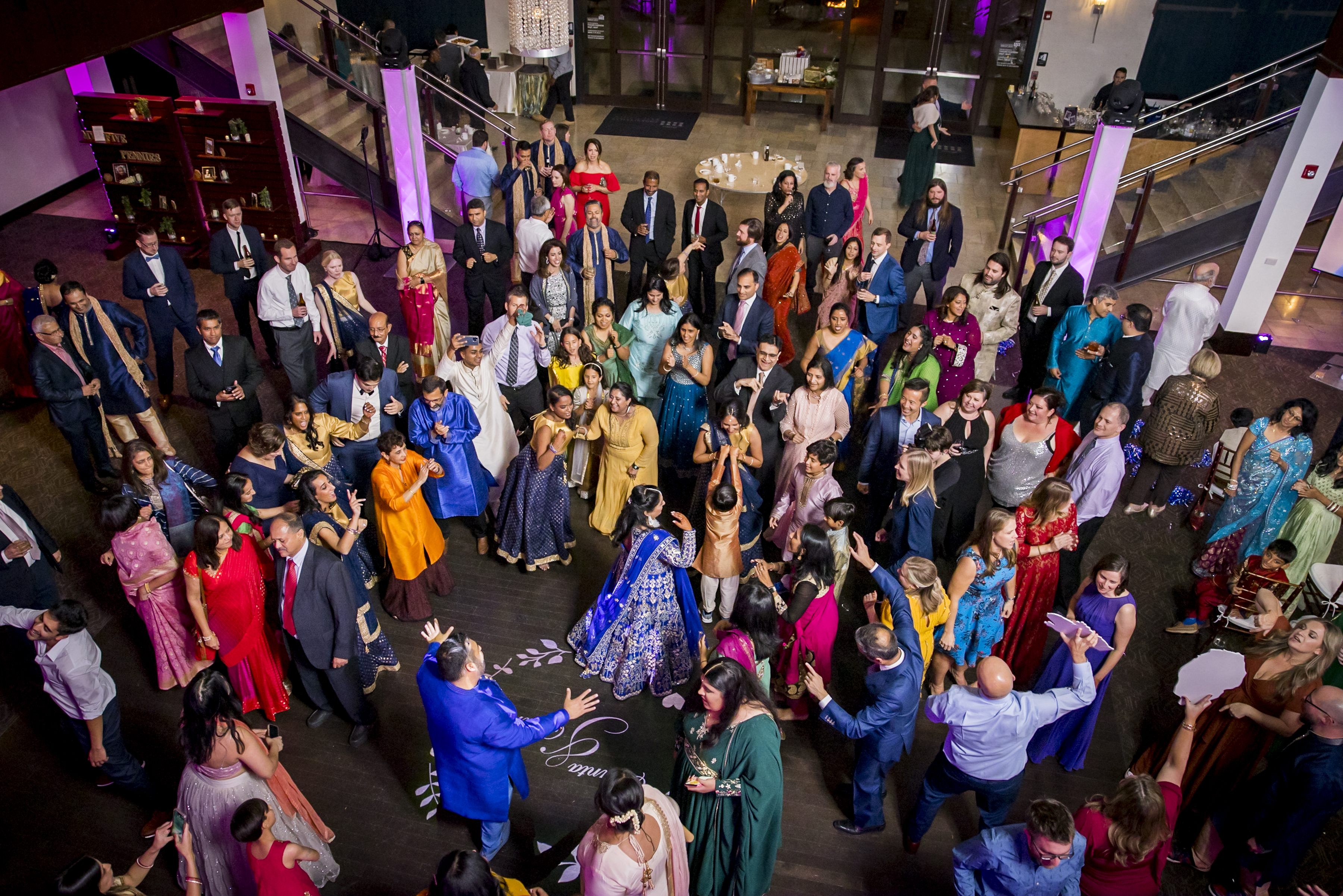 A large group of people in colorful attire dance and celebrate in a decorated indoor venue.