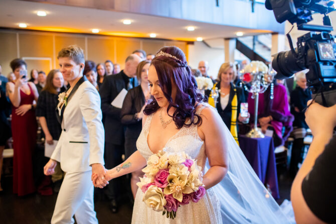 A bride and groom walk down the aisle, holding hands, surrounded by wedding guests and photographers.