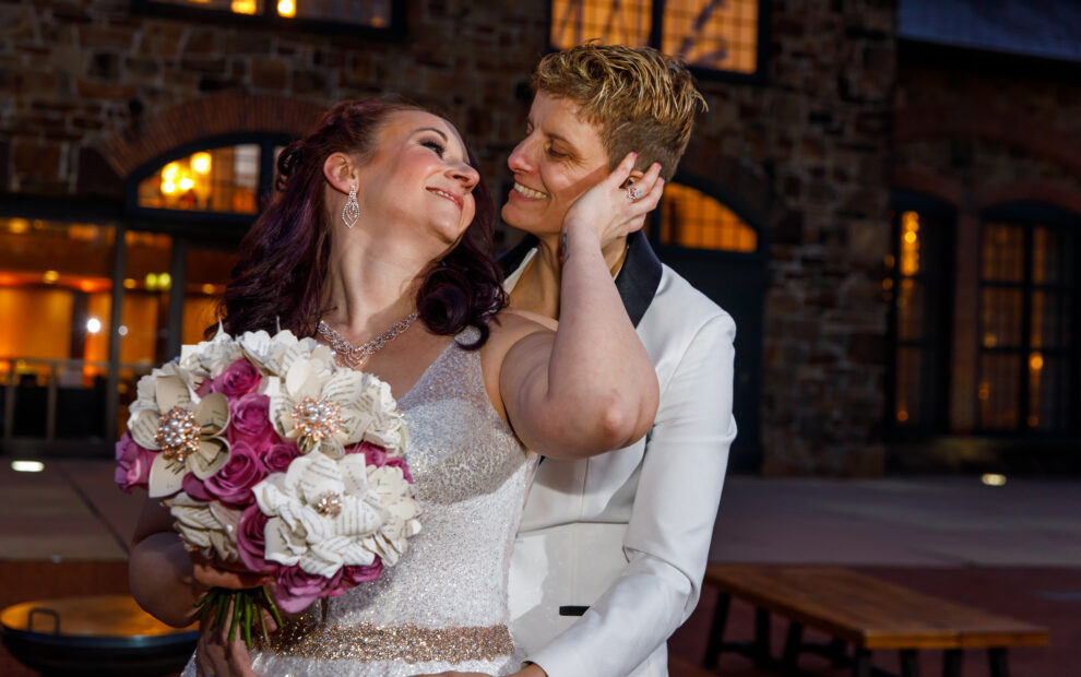 A happy couple in wedding attire embrace and smile at each other, holding a bouquet of pink and white flowers.