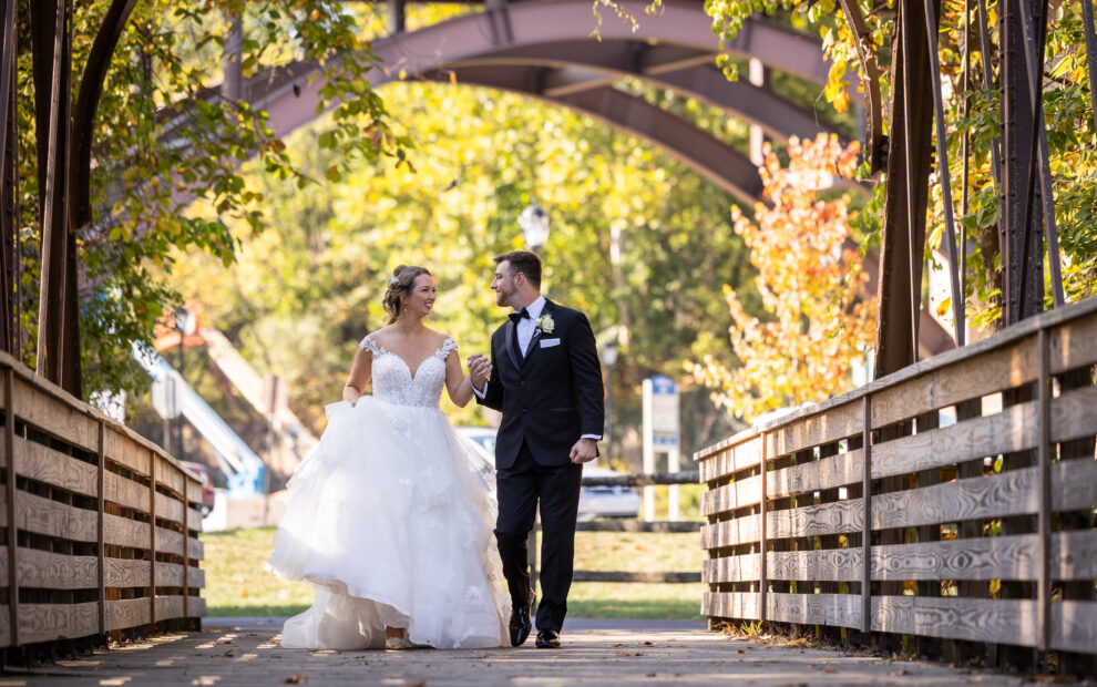 Bride and groom holding hands, walking on a wooden bridge outdoors under an arched structure.