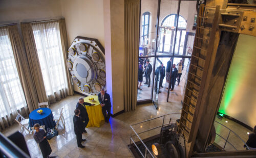 People in formal attire mingle at an indoor event space with tall windows and industrial decor.