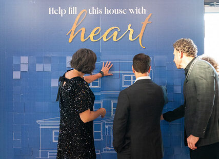 Three people in formal attire add squares to a blue board that reads, "Help fill this house with heart.
