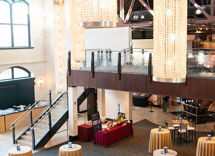 Spacious event hall with tall chandeliers, round tables with gold cloths, and a mezzanine level.