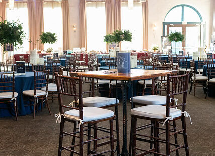 A banquet hall with tall tables, chairs, blue tablecloths, and large windows letting in natural light.