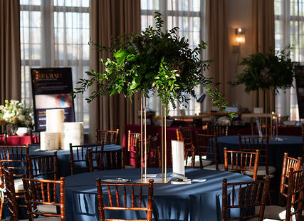 Round table with blue tablecloth, wood chairs, and a tall green centerpiece in a sunlit event hall.