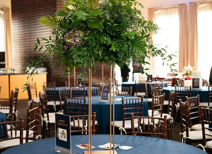 Tall green floral centerpiece on a round table with a blue tablecloth in an event venue with chairs.