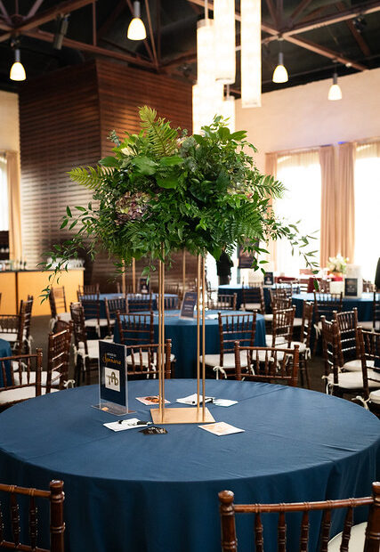 Tall green floral centerpiece on a round table with a blue tablecloth in an event venue with chairs.