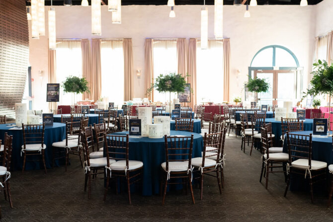 Event hall with round tables covered in blue cloth, brown chairs, and large windows letting in natural light.